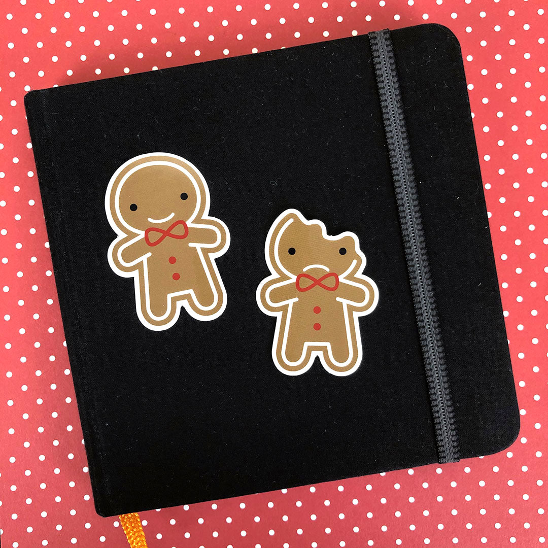 Two die cut vinyl stickers of happy and sad bitten gingerbread men, displayed on a small sketchbook