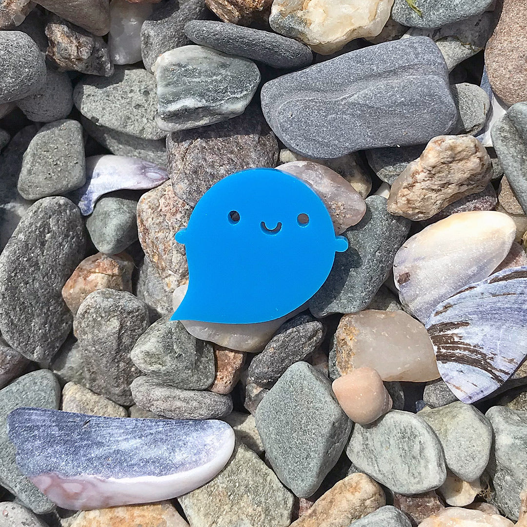 The blue Rainbow Ghost brooch sitting on a pile of beach pebbles
