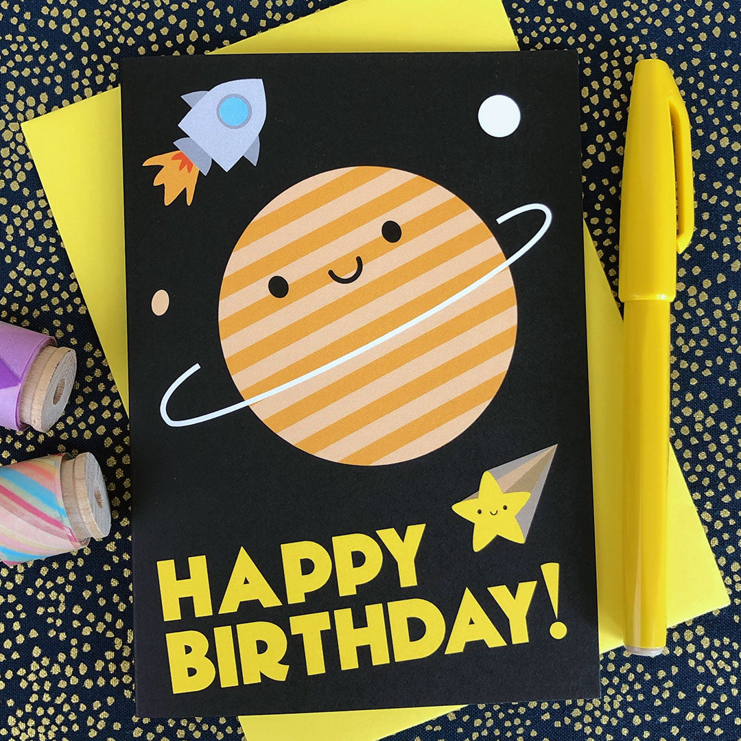 An illustrated card with a happy ringed planet, rocket and shooting star plus the words 'happy birthday!'
