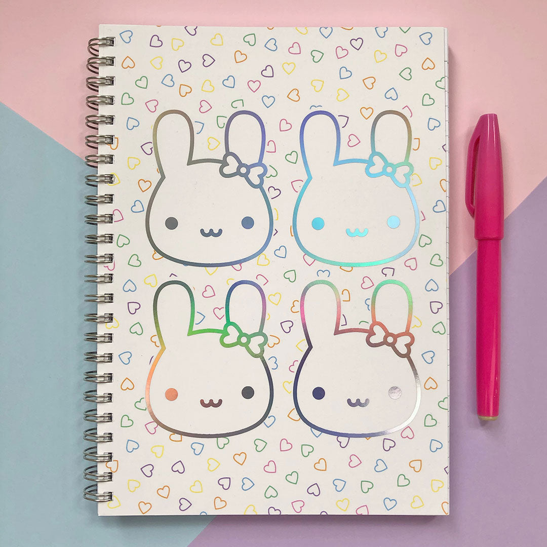 Spiral bound notebook with 4 kawaii bunny faces in rainbow holographic foil on a background of small scattered hearts