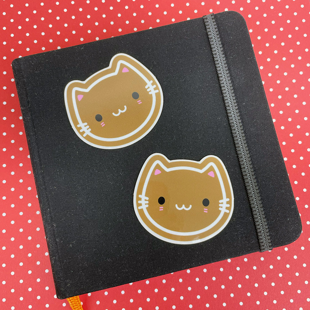 Two die cut vinyl stickers of iced gingerbread cookie cats, displayed on a small sketchbook