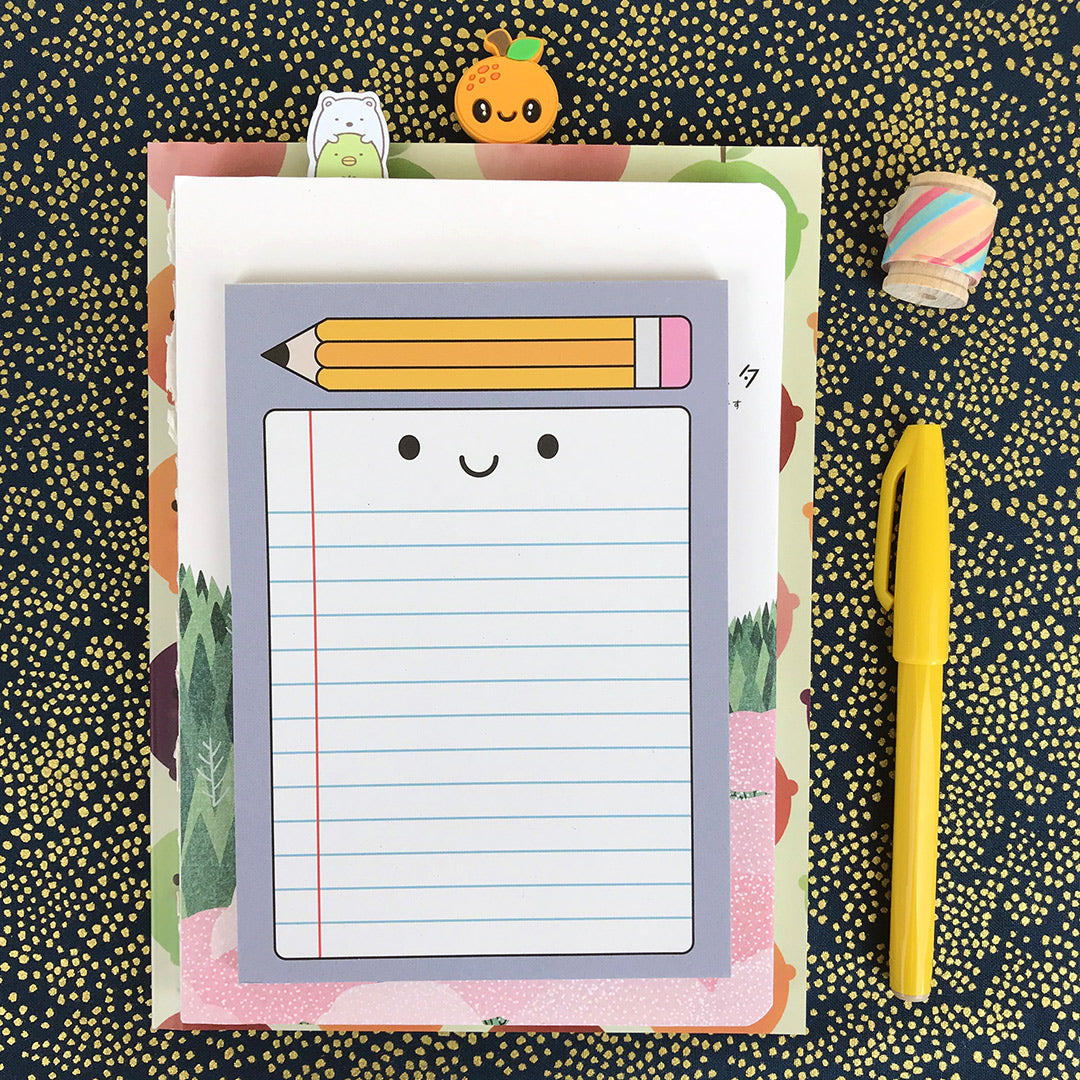 A notepad with an illustrated design of a happy sheet of lined paper and pencil on top, displayed on a pile of notebooks with pen and washi tape